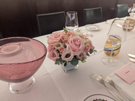 A pink floral arrangement with roses on a dining table set with glassware.
