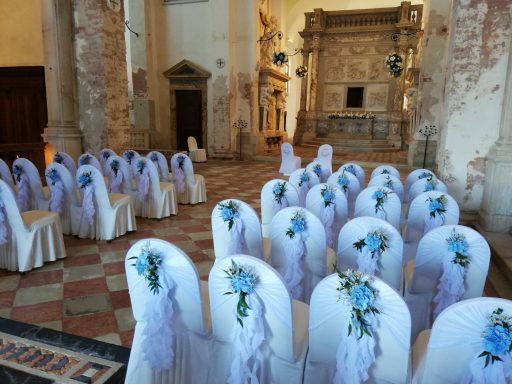 Sala decorata per un evento, con sedie bianche e nastri blu, in un ambiente elegante.