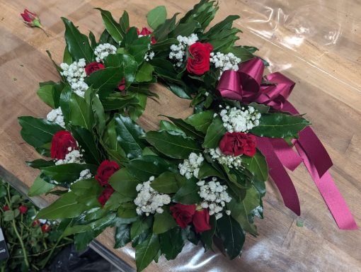 Corona di fiori con rose rosse e del foliage, decorata con un nastro bordeaux.