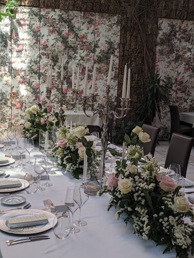 Tavola elegantemente apparecchiata con fiori e candelabri, decorazioni floreali sullo sfondo.