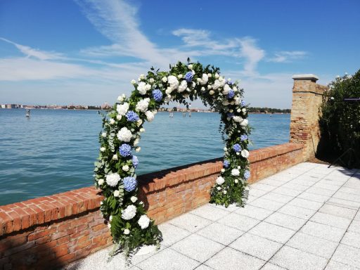 Arcata decorata con fiori bianchi e blu, affacciata su un lago sotto un cielo sereno.