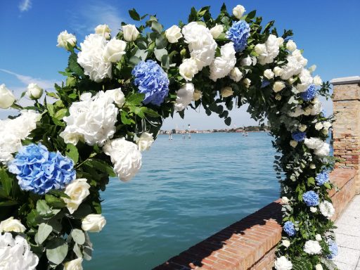 Arco di fiori bianchi e blu con vista su acqua e cielo sereno.