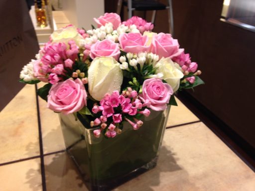 Bouquet of pink and white roses with delicate white flowers in a square vase.